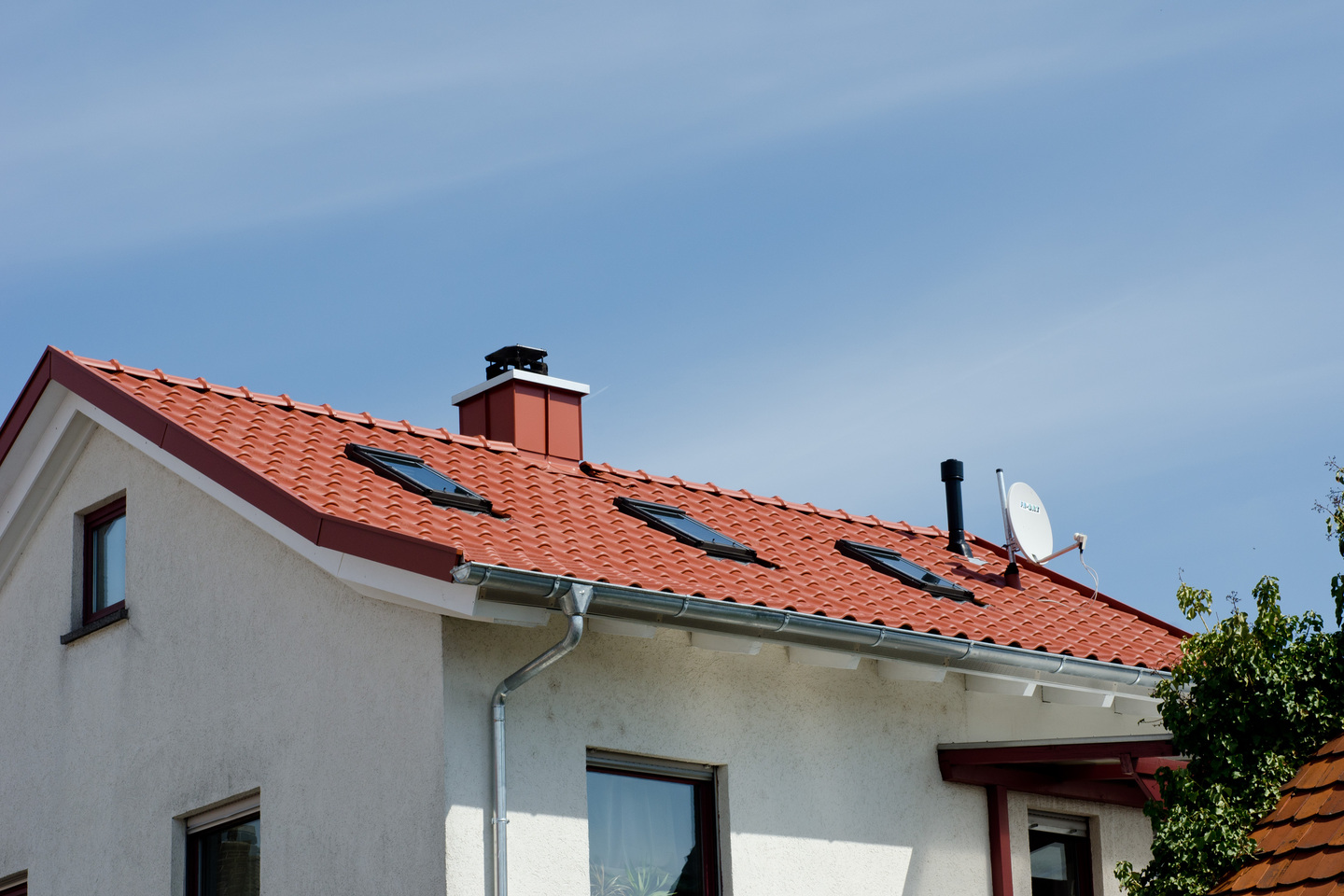 Dach eines Hauses mit roten Ziegeln, Dachfenstern, Schornstein und Satellitenschüssel unter blauem Himmel.
