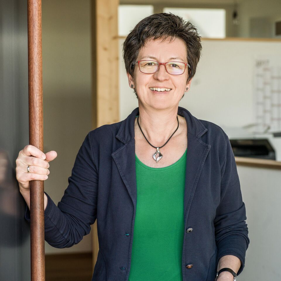 Eine lächelnde Person mit kurzen Haaren und Brille hält einen Holzstab und trägt ein grünes Oberteil unter einer blauen Jacke.