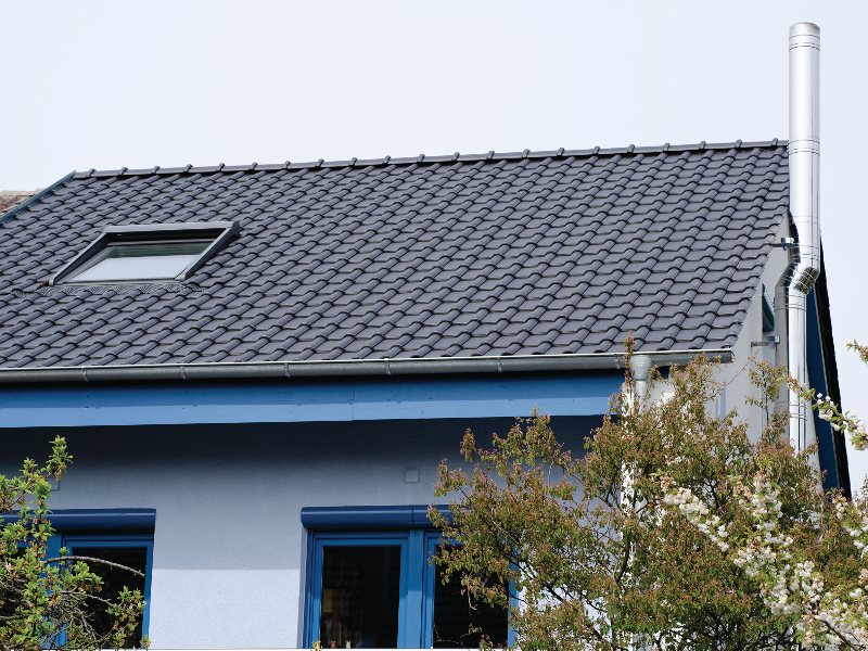 Ein modernes Haus mit blauen Fensterrahmen, dunklem Dach und einem Schornstein, umgeben von Bäumen.