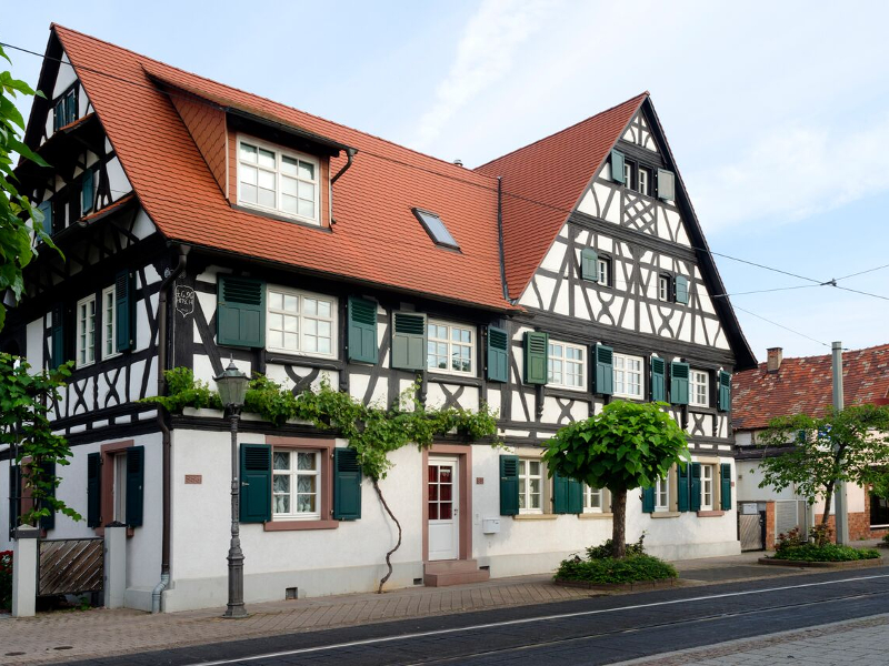 Ein traditionelles Fachwerkhaus mit rotem Dach, weißen Wänden und grünen Fensterläden steht an einer Straße mit Bäumen und Laternen.