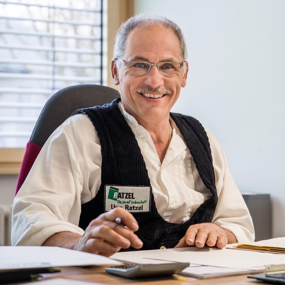 Ein lächelnder Mann mit Brille und Namensschild sitzt an einem Schreibtisch in einem Büro.