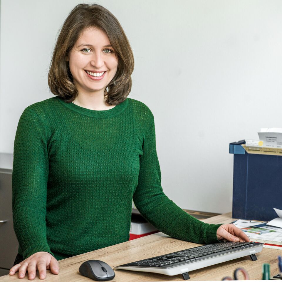 Eine Frau im grünen Pullover sitzt lächelnd an einem Schreibtisch mit Tastatur und Maus.