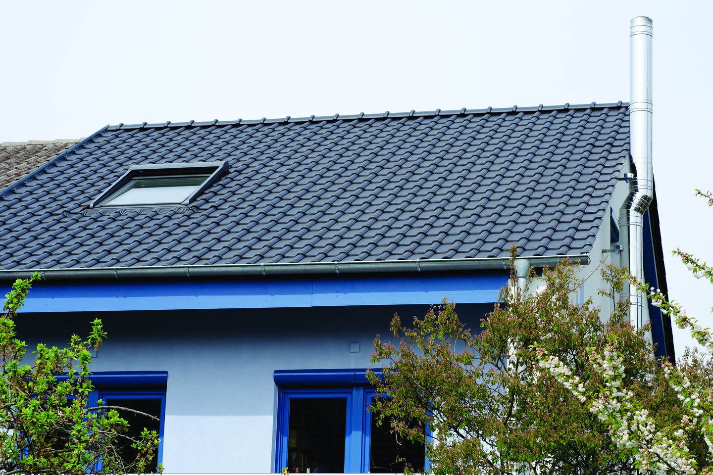 Ein Haus mit blauem Anstrich, grauem Dach und einem Dachfenster, umgeben von Bäumen.