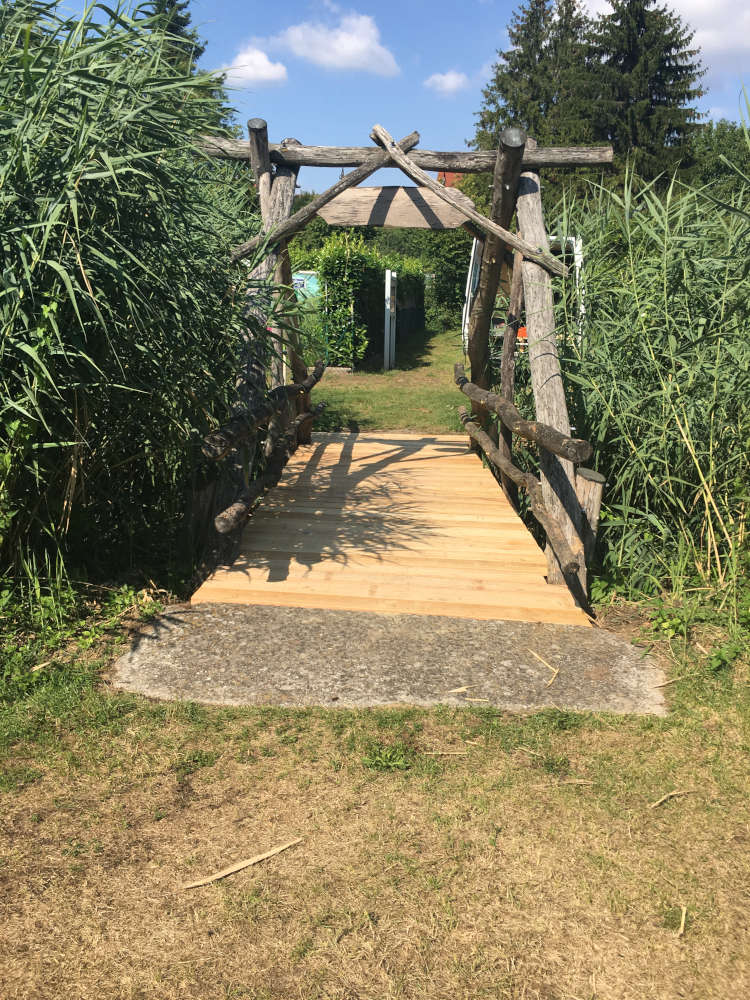 Eine hölzerne Brücke führt über eine Grasfläche zu einem Torbogen, umgeben von hohem Gras und Bäumen unter blauem Himmel.
