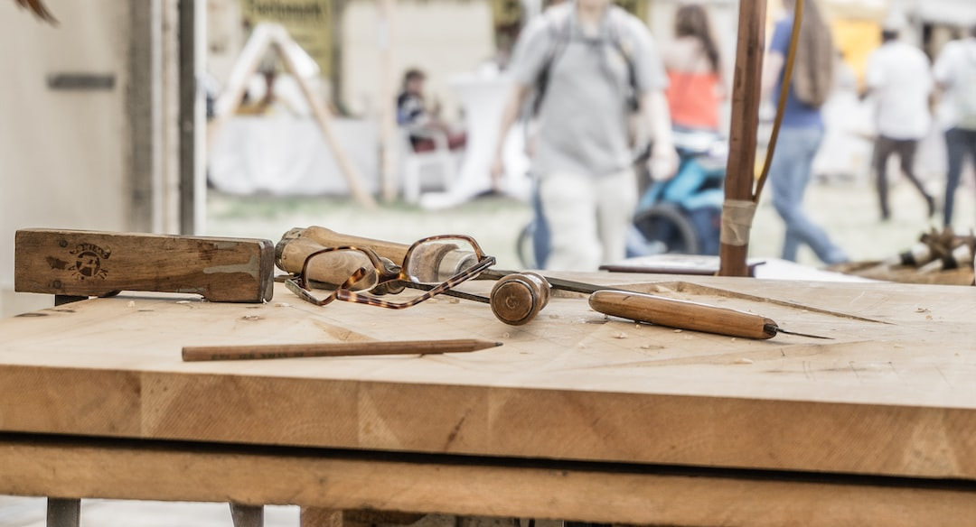 Holzwerkzeuge, darunter ein Hammer, eine Zange und ein Bleistift, liegen auf einem Holztisch, im Hintergrund sind Menschen auf einem Markt zu sehen.